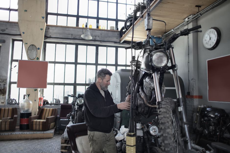 Side view of bearded adult mechanic in casual uniform standing near motorcycle and working with engine in workshop
