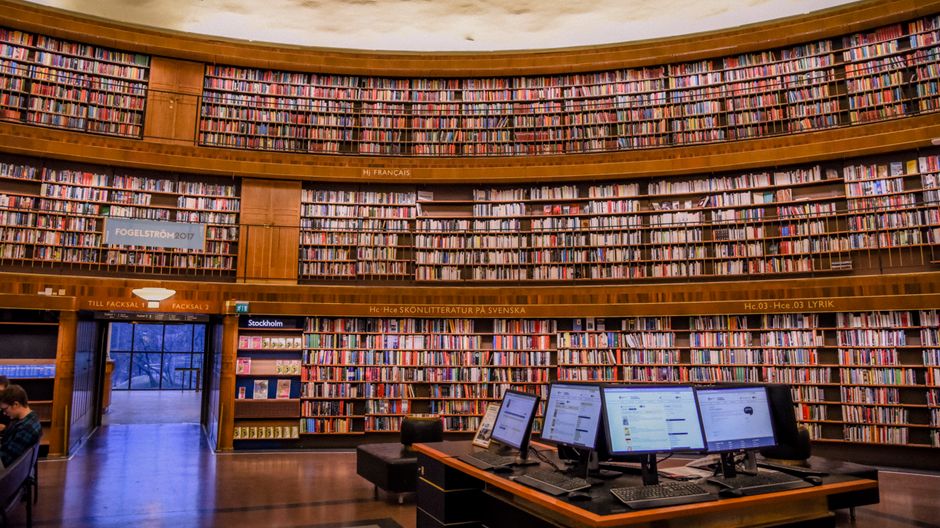 Public library with collection of colorful books on wooden bookshelves near table with computer monitors