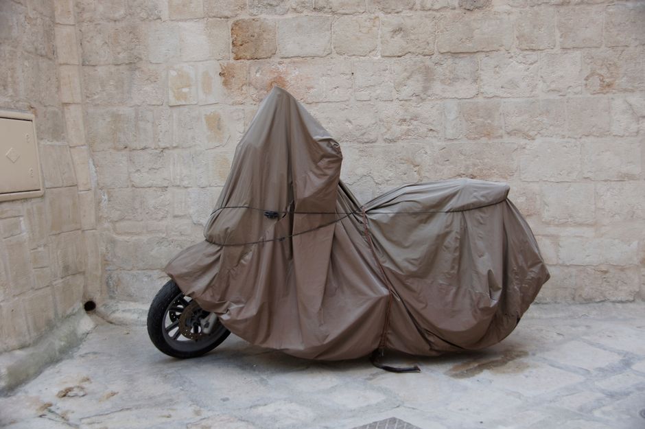 A motorcycle covered with a tarp is parked against a stone wall in an urban environment.